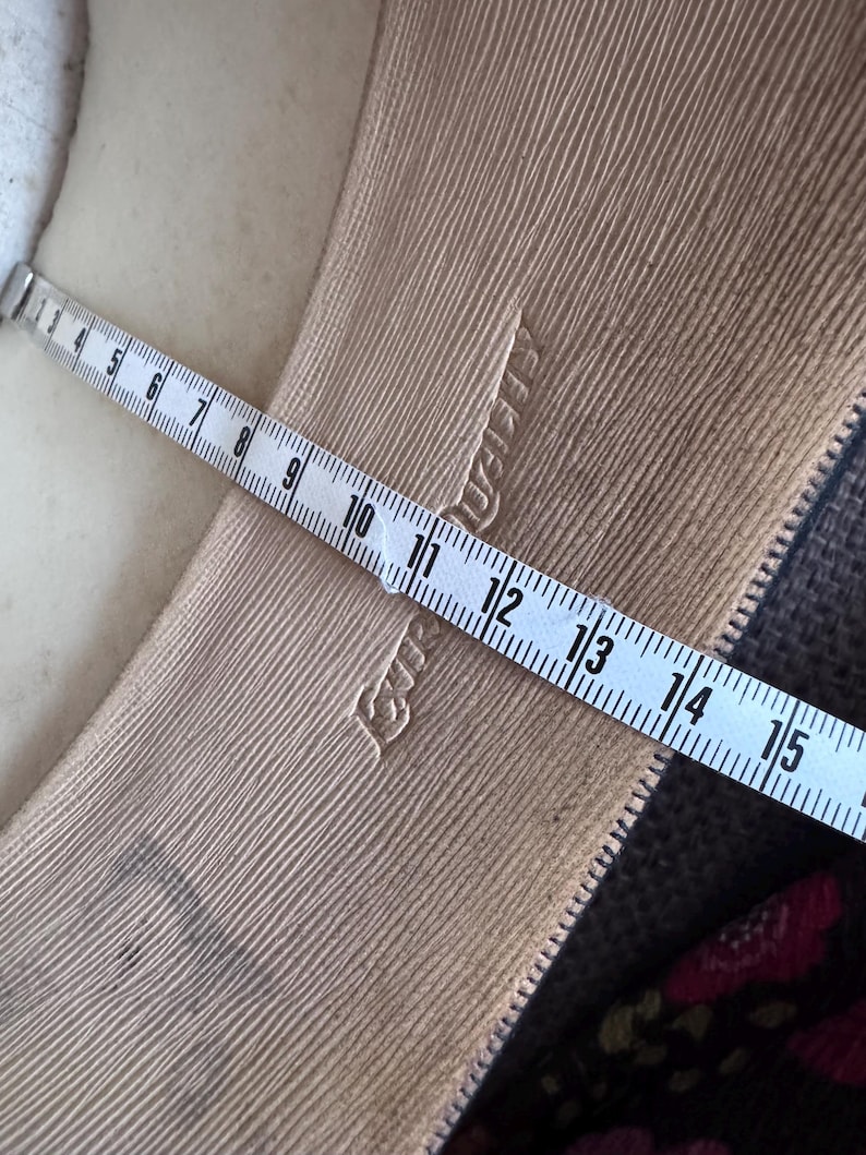 May include: Close-up of a beige fabric with a textured, ribbed pattern. A white measuring tape is laid across the fabric, showing measurements in inches. The fabric has a subtle sheen and a zipper is visible.