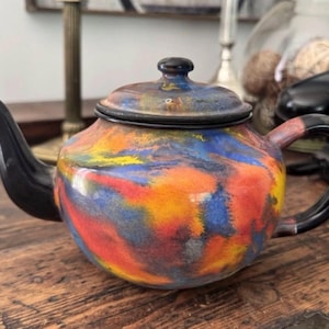 May include: A round teapot featuring a vibrant, multi-coloured abstract glaze in shades of orange, blue, yellow, and red. The teapot includes a matching lid, a solid black curved handle, and a matching black spout, creating a bold contrast.