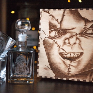 May include: A wooden box featuring a burned image of a scary doll's face. The box is part of a liquor set that includes a glass decanter and two shot glasses, all with etched images of the same doll.