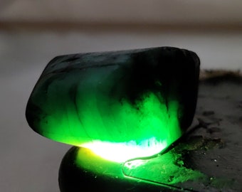 May include: A dark green gemstone with a light green interior, illuminated from below. The gemstone is resting on a black surface.