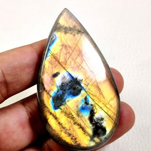 Peut inclure: Une pierre précieuse de labradorite en forme de goutte polie. La pierre présente un mélange de couleurs, notamment du jaune, du marron, du bleu et du noir. La pierre est tenue dans une main, mettant en valeur sa taille et sa forme.