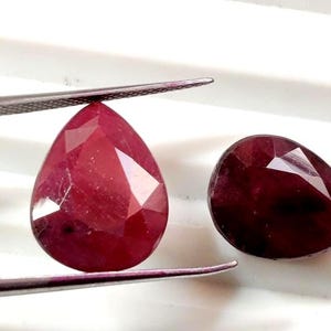 Può includere: Due rubini sfaccettati a forma di pera. Uno è tenuto da una pinzetta, che mostra il suo colore rosso intenso e le sfaccettature tagliate. L'altro rubino, anch'esso a forma di pera, è accanto, mostrando una tonalità ricca e una superficie lucida simile.