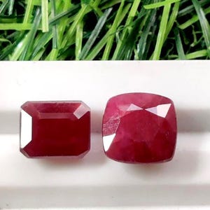 May include: Two faceted, deep red gemstones. One is rectangular with cut corners, the other is a cushion cut. The gems are displayed on a white surface, with a background of green artificial grass.