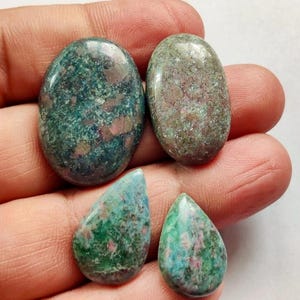 Peut inclure: Quatre pierres précieuses polies dans les tons de vert, de bleu et de rose. Deux sont de forme ovale et deux en forme de goutte. Les pierres ont un aspect moucheté, avec un mélange de couleurs et de textures. Les pierres sont tenues dans une main.