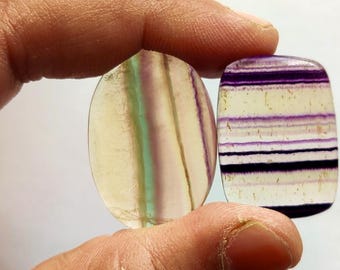 May include: Two oval-shaped fluorite cabochons, one with a light green and purple striped pattern and the other with a white and purple striped pattern.