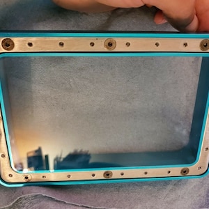 May include: A rectangular frame with a clear glass center. The frame has a bright teal border and a silver metal trim secured with small screws. The glass reflects a cityscape. The frame is held by a hand.