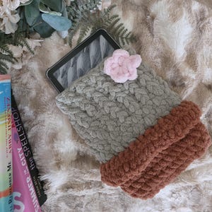 Peut inclure: Une housse de tablette tricotée à la main, en fil vert sauge et rouille, ornée d'une fleur rose. La housse repose sur une couverture en fausse fourrure moelleuse, avec un livre et une composition florale en arrière-plan.