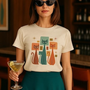 Könnte beinhalten: Cremefarbenes T-Shirt mit einem Retro-Design von drei stilisierten Katzen in Braun, Türkis und Orange. Die Katzen sind vor einem Hintergrund aus geometrischen Formen und Sternen platziert. Die Person trägt eine schwarze Sonnenbrille und einen türkisfarbenen Rock.