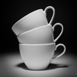 May include: Three white ceramic coffee mugs stacked on top of each other. The mugs are all the same size and shape, and they have a simple, elegant design.