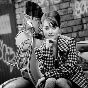 Peut inclure: Une photo en noir et blanc d'une femme portant une veste à carreaux assise sur un scooter vintage. Le scooter a un porte-bagages chromé et un phare chromé. La femme regarde la caméra et a la main sur le menton.