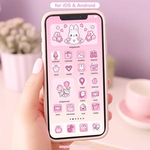 May include: A pink and white smartphone with a pink and white bunny rabbit icon on the home screen. The home screen is filled with pink and white icons for apps such as calendar, weather, mail, camera, maps, internet, bank, calculator, and twitter. The text "widgetsmith" appears on the screen. The phone is held in a hand.
