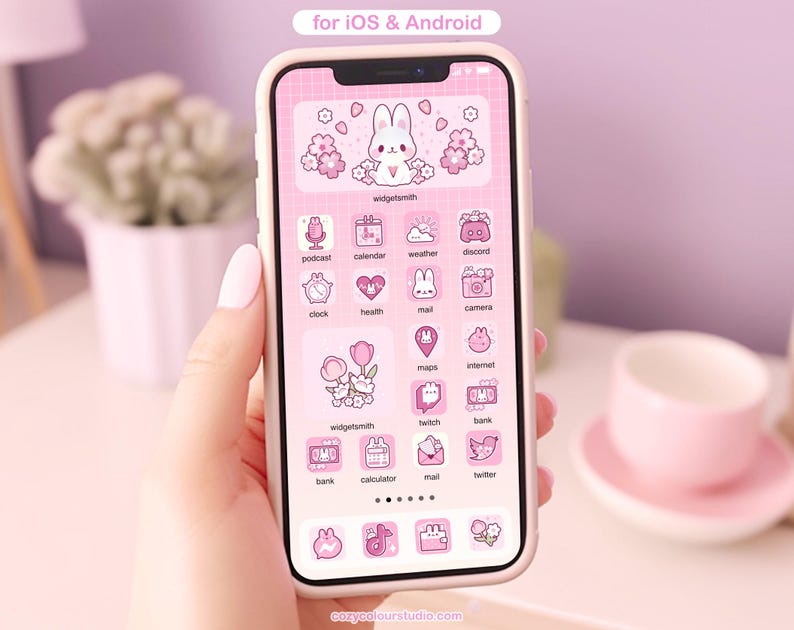 May include: A pink and white smartphone with a pink and white bunny rabbit icon on the home screen. The home screen is filled with pink and white icons for apps such as calendar, weather, mail, camera, maps, internet, bank, calculator, and twitter. The text "widgetsmith" appears on the screen. The phone is held in a hand.