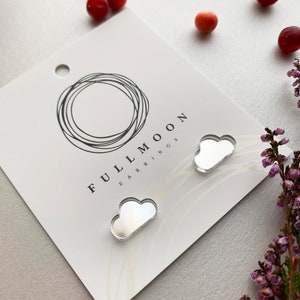 May include: Cloud-shaped earrings on a white card with the text "FULLMOON EARRINGS". The earrings are silver and reflective. The card has a circular design at the top. Small red berries and purple flowers are scattered around the card.