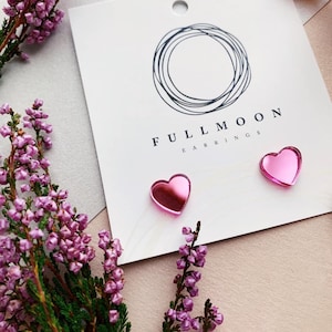 May include: A pair of pink heart-shaped earrings with a mirrored finish. The earrings are displayed on a white card with the text "FULL MOON EARRINGS".
