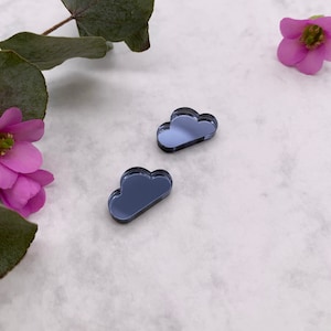 Puede incluir: Dos dijes espejados en forma de nube de color azul oscuro. Los dijes son reflectantes y tienen una superficie lisa. El fondo es blanco con flores rosas y hojas verdes.