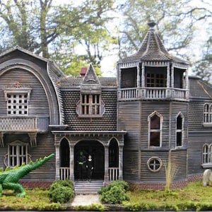 May include: A model of a spooky, gray, Victorian-style house with a turret and a dragon figurine in the front yard. The house has a porch with a black door and a small, green bush in front of it.