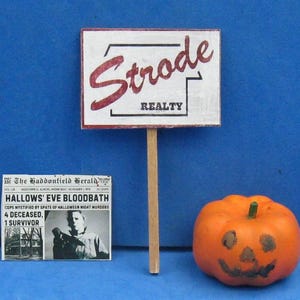 May include: A miniature Halloween-themed scene featuring a Strode Realty sign, a newspaper with the headline "HALLOWS' EVE BLOODBATH," and a carved pumpkin. The sign has red text on a white background. The pumpkin is orange with a carved face.