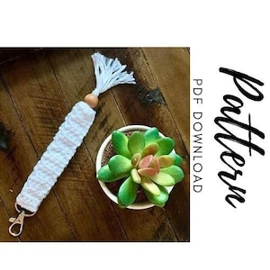 May include: White crocheted keychain with a wooden bead and a tassel. The keychain is attached to a silver key ring. The keychain is on a wooden surface with a small green succulent plant in a white pot.