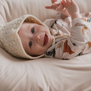 May include: A baby wearing a white crocheted bonnet and a white and brown long-sleeved onesie with a woodland print. The baby is lying on a cream-colored blanket and smiling.