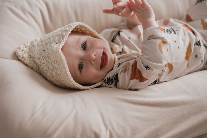 May include: A baby wearing a white crocheted bonnet and a white and brown long-sleeved onesie with a woodland print. The baby is lying on a cream-colored blanket and smiling.