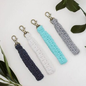 May include: Four crocheted keychains in different colours: grey, white, light blue, and dark blue. Each keychain has a gold metal clasp.
