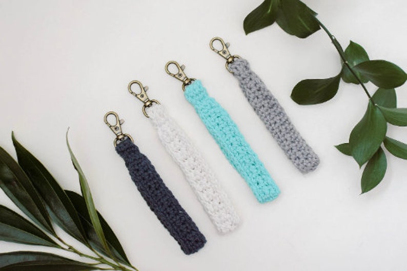 May include: Four crocheted keychains in different colours: grey, white, light blue, and dark blue. Each keychain has a gold metal clasp.