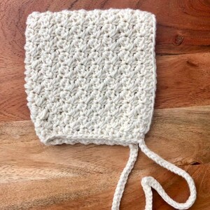 May include: A white crocheted baby bonnet with a tie string. The bonnet is made with a simple stitch pattern and has a soft, delicate look.