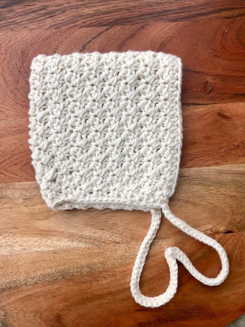 May include: A white crocheted baby bonnet with a tie string. The bonnet is made with a simple stitch pattern and has a soft, delicate look.