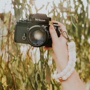 May include: A black Nikon camera with a white crocheted camera strap. The camera is being held by a person's hand.