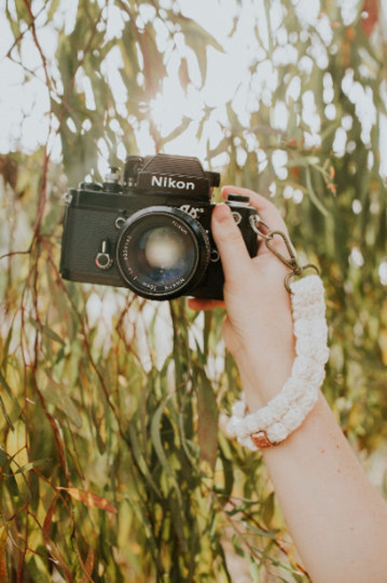May include: A black Nikon camera with a white crocheted camera strap. The camera is being held by a person's hand.