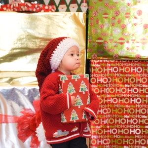 以下が含まれることがあります： 赤と白のクロシェのサンタ帽をかぶった小さな子供が、クリスマスツリーのデザインが施された赤と緑の包装紙で包まれたプレゼントボックスを持っています。子供は、包装されたプレゼントで飾られた壁の前に立っています。