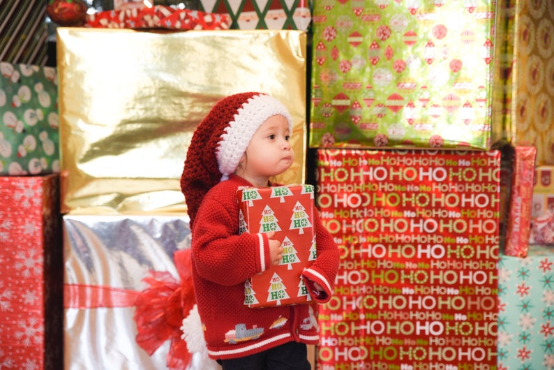 以下が含まれることがあります： 赤と白のクロシェのサンタ帽をかぶった小さな子供が、クリスマスツリーのデザインが施された赤と緑の包装紙で包まれたプレゼントボックスを持っています。子供は、包装されたプレゼントで飾られた壁の前に立っています。
