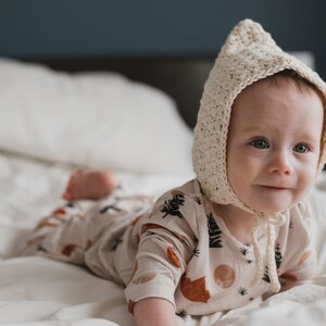 May include: A baby wearing a cream crocheted bonnet and a cream and brown patterned onesie lies on a white bed. The baby is looking to the right of the frame.