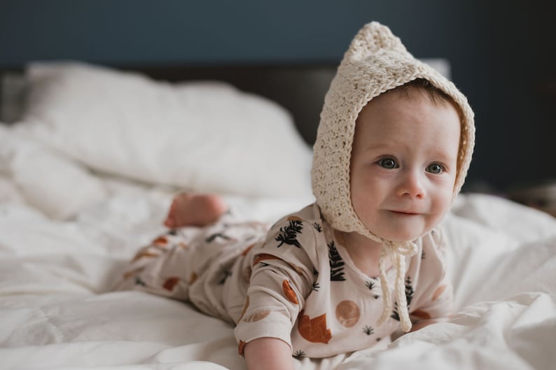 May include: A baby wearing a cream crocheted bonnet and a cream and brown patterned onesie lies on a white bed. The baby is looking to the right of the frame.