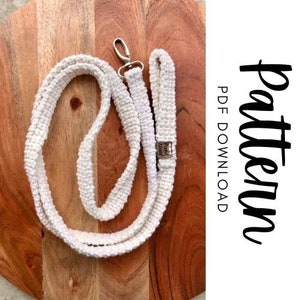 Op de afbeelding: Witte gecrochete hondenriem met een zilveren clip. De riem ligt op een houten oppervlak. De tekst "Pattern PDF Download" is zichtbaar in de afbeelding.