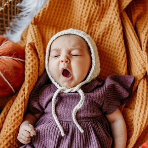 May include: A baby wearing a purple dress and a white crocheted bonnet is lying in a woven basket with a yellow blanket. The baby is yawning and there are several small pumpkins around the baby.