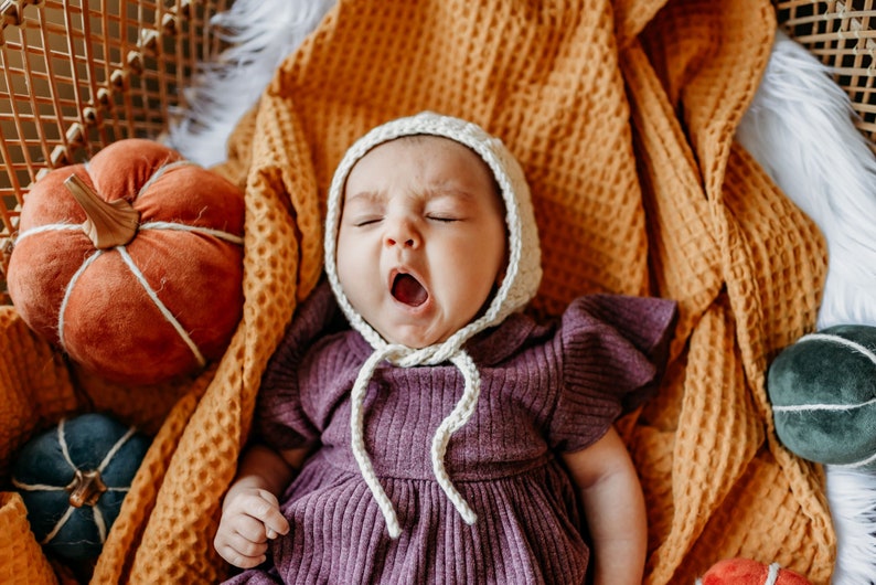 May include: A baby wearing a purple dress and a white crocheted bonnet is lying in a woven basket with a yellow blanket. The baby is yawning and there are several small pumpkins around the baby.