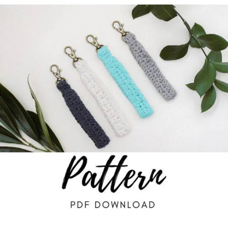 May include: Five crocheted keychains in different colours, including grey, white, and turquoise, with gold key rings. The keychains are arranged on a white background with a green leafy branch. The text "Pattern PDF Download" is at the bottom of the image.