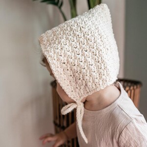 May include: A white crocheted bonnet with a tie at the back. The bonnet is worn by a child wearing a light pink dress.