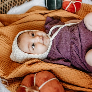 May include: A baby wearing a white crocheted bonnet and a purple knitted onesie is lying on a yellow blanket with three velvet pumpkins. The pumpkins are green, red, and orange.