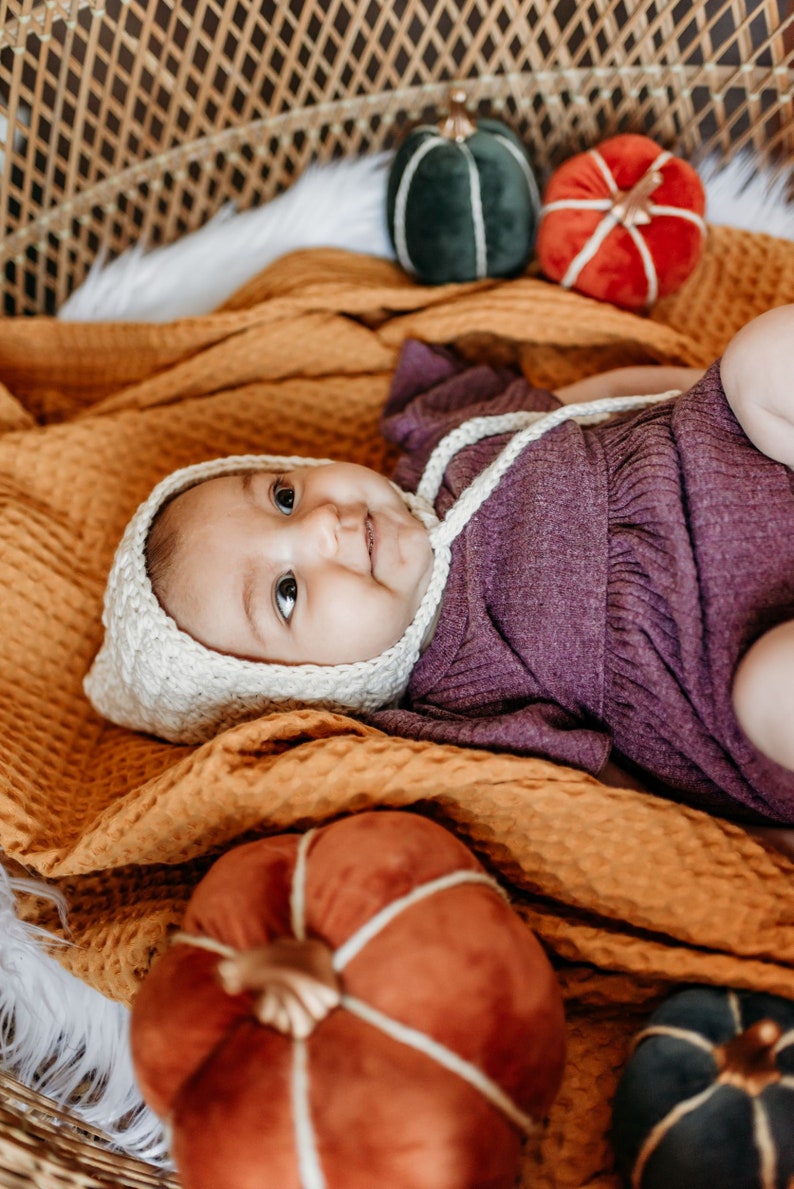 May include: A baby wearing a white crocheted bonnet and a purple knitted onesie is lying on a yellow blanket with three velvet pumpkins. The pumpkins are green, red, and orange.