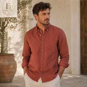 May include: A long-sleeved button-down shirt in a solid terracotta orange color. The shirt features a classic collar, a single chest pocket on the left side, and light-colored buttons running down the center placket. The fabric has a visible texture.