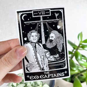 Puede incluir: Adhesivo en blanco y negro con dos hombres vestidos con ropa histórica, con el texto "The Co-Captains". El diseño incluye un mástil de barco, una luna creciente y el número romano VIII. El adhesivo se sostiene sobre un fondo de follaje verde.