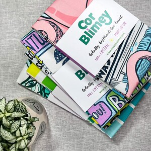 Może przedstawiać: Stos czterech kolorowych ręcznik&oacute;w kuchennych z r&oacute;żnymi wzorami graficznymi, w tym r&oacute;żowym flamingiem i tekstem. Każdy ręcznik jest oznaczony napisem "Cor Blimey" i "totally brilliant tea towel". Ręczniki wykonane są w 100% z bawełny.
