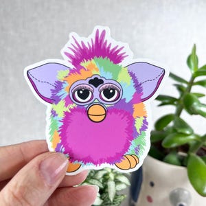 Könnte beinhalten: Ein farbenfroher Aufkleber mit einer furby-ähnlichen Kreatur. Der Aufkleber hat eine lebendige Mischung aus Farben, darunter Lila, Rosa, Grün und Orange. Er hat große, ausdrucksstarke Augen, einen gelben Schnabel und lila Ohren. Der Aufkleber wird von einer Hand gehalten.