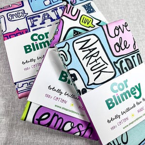 May include: Four folded tea towels with colorful graphic designs and text, including the words "Grotty," "Narful," and "Love." Each towel is wrapped with a white band that reads "Cor Blimey" and "totally brilliant tea towels."