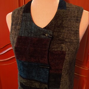May include: A patchwork vest with a V-neck and two toggle closures. The vest features a mix of textured fabrics in shades of gray, blue, and burgundy. The collar is a dark blue. The vest is displayed on a mannequin.