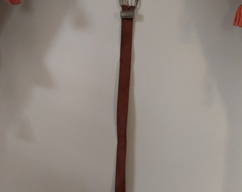 May include: A brown leather belt with a silver-colored buckle. The belt is long and slender, with a simple design. The buckle has a decorative edge. The belt is hanging against a white wall.