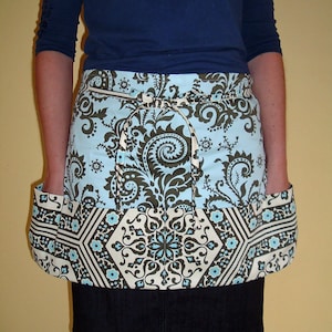 May include: A blue and white patterned apron with a brown floral design. The apron has two large pockets on the sides and a tie at the waist.