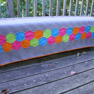 Pode incluir: Um corredor de mesa acolchoado cinza com uma borda de tecido laranja. O corredor de mesa tem uma fileira de remendos de tecido hexagonais coloridos em tons de rosa, verde, azul e laranja.